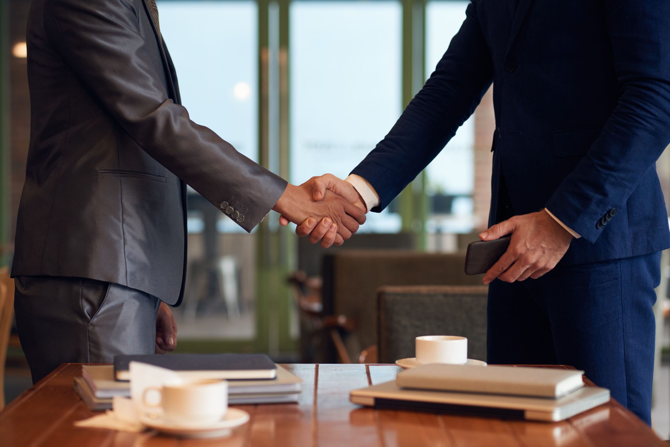 Two people in business attire are shaking hands across a table with documents, notebooks, and coffee cups in an office setting.