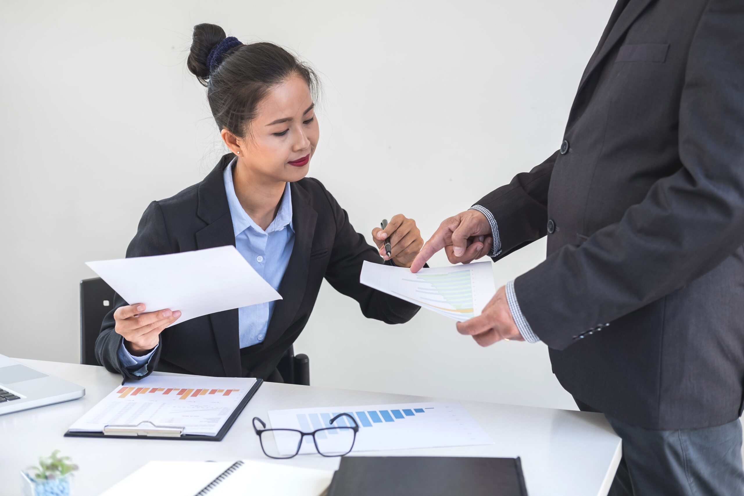 Two business professionals review and discuss printed charts and documents at a desk with a laptop, papers, and glasses.