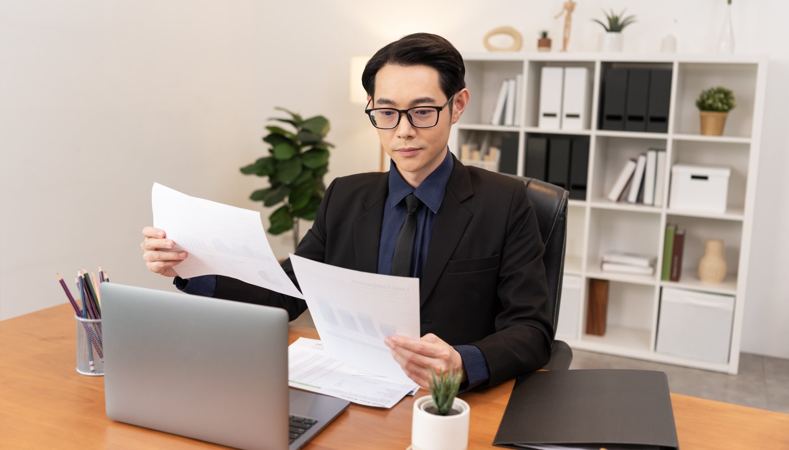 A man in a suit sits at a desk in an office, examining documents in his hands with a laptop, plant, and folders on the desk, and shelves in the background.
