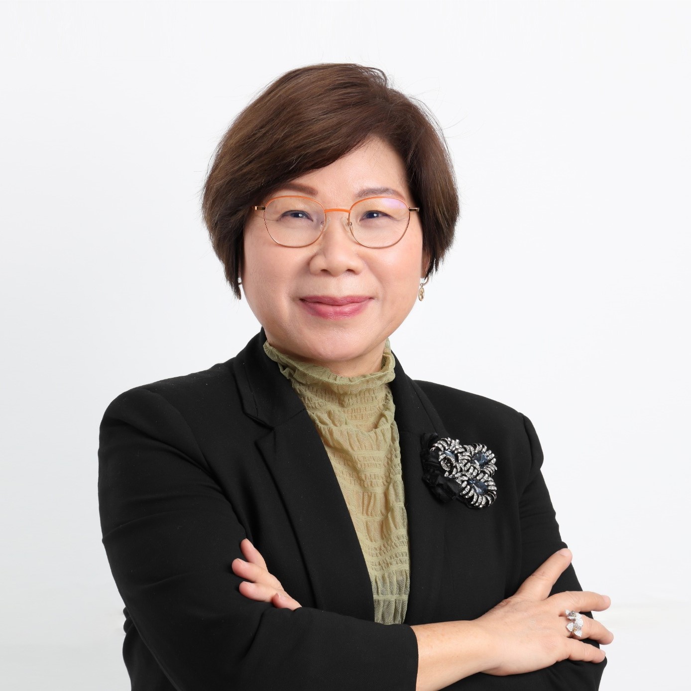 Woman with short brown hair, glasses, and a black blazer with a brooch stands with arms crossed, smiling in front of a plain white background.