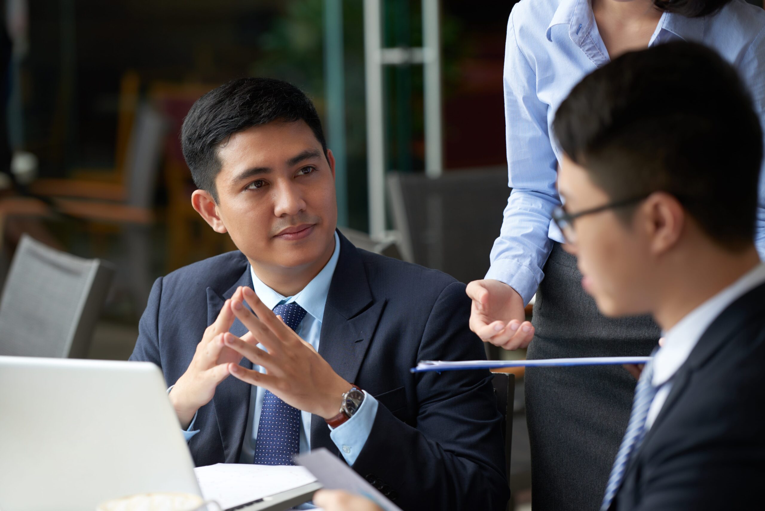 Three people in business attire have a discussion around a table with a laptop and documents in a professional office setting.