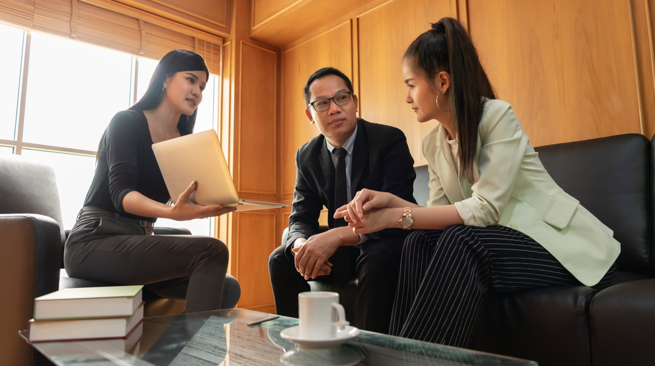 Three people in business attire sit and discuss something in an office setting; one holds a laptop, and a coffee cup sits on the glass table in front of them.