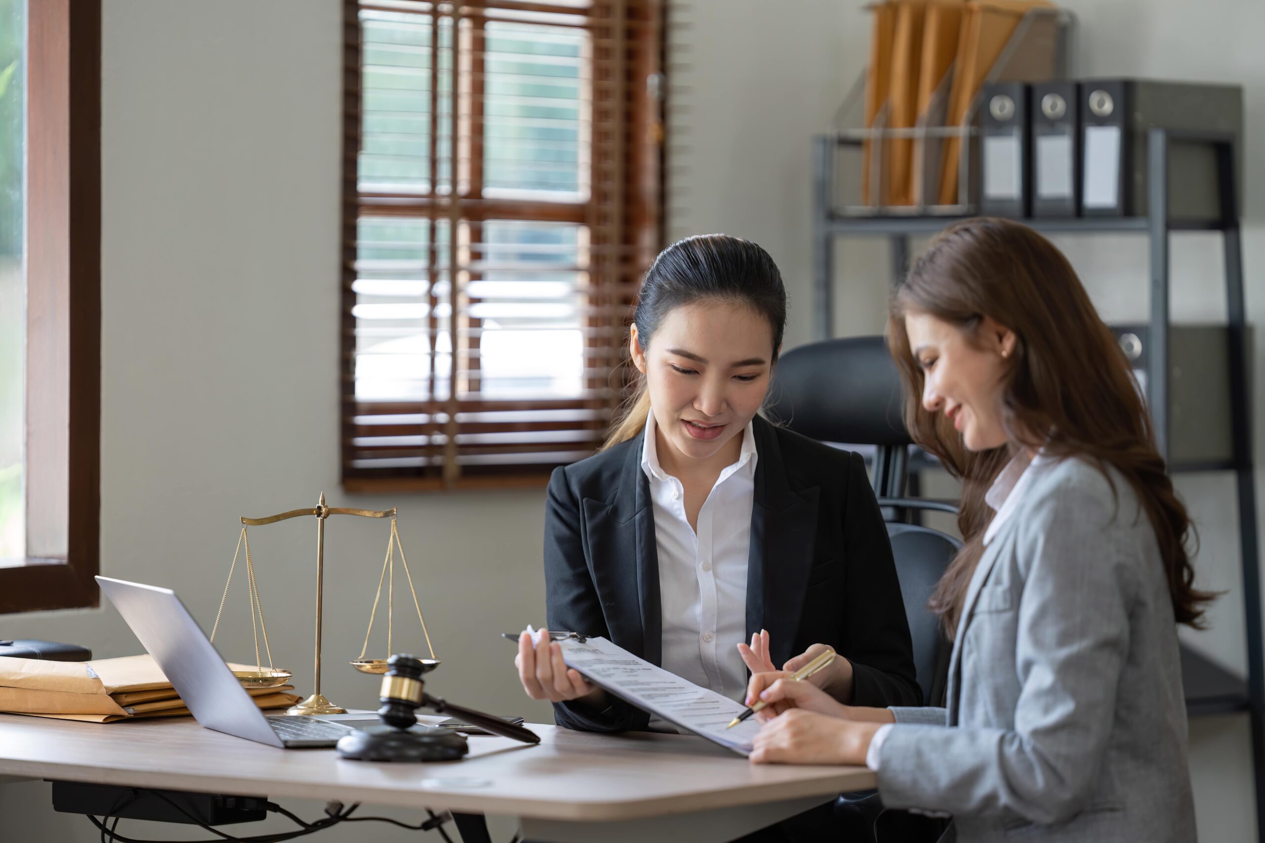 Two women in business attire sit at a desk with legal documents, a gavel, and scales of justice, discussing paperwork in an office setting.