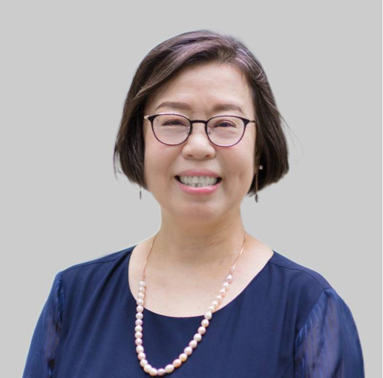 Woman with short dark hair, glasses, and a pearl necklace smiling and wearing a navy blue blouse against a plain gray background.