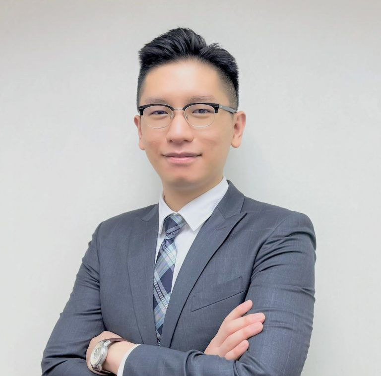 A man wearing glasses and a gray suit with a plaid tie, standing with arms crossed against a plain light background.